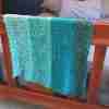 Ocean Layers Wrap Crochet Pattern by ReVeDesignCo - hanging at the end of a bed - in shades of teal